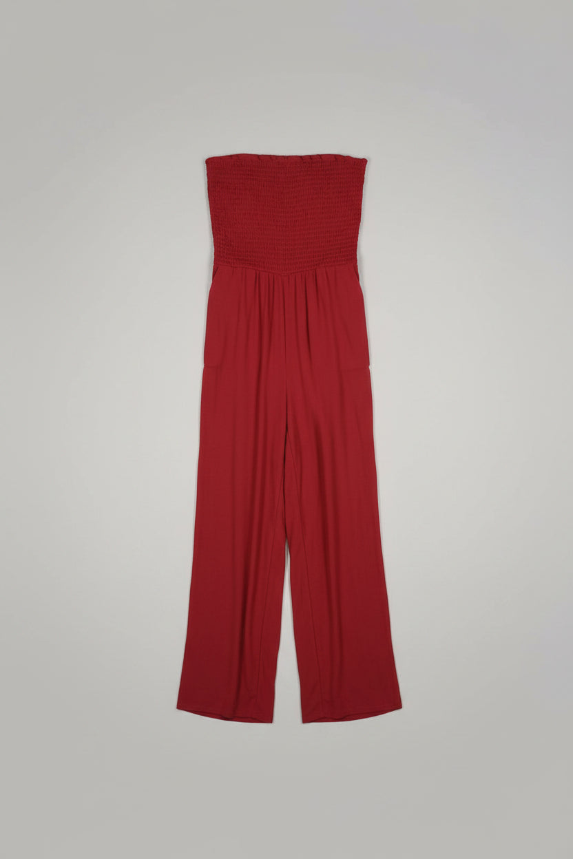 A bright red sleeveless smocked bodice dress or jumpsuit being held up by a hand, shown against an indoor background with a white door and window.