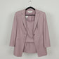 A dusty pink three-piece suit set consisting of a blazer, vest, and matching skirt, held up on a hanger by a person's hand in front of a wooden floor background.