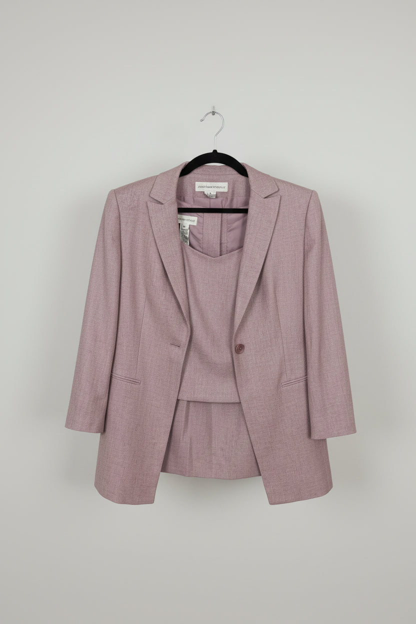 A dusty pink three-piece suit set consisting of a blazer, vest, and matching skirt, held up on a hanger by a person's hand in front of a wooden floor background.