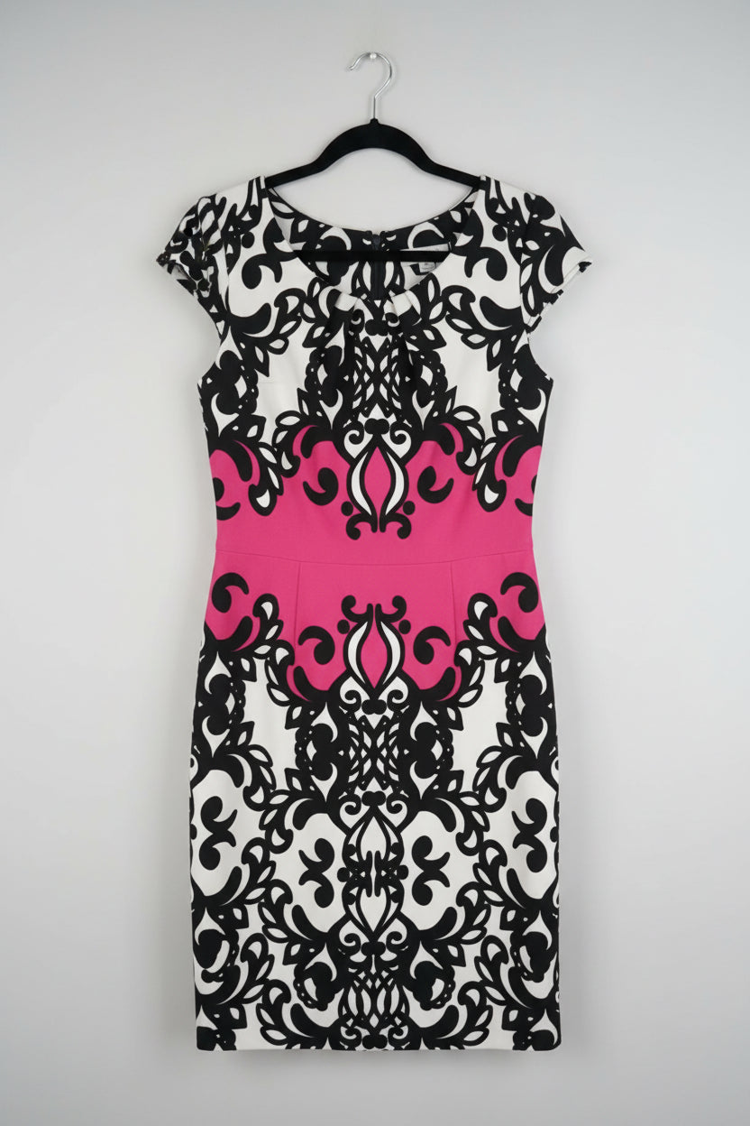 A sleeveless sheath dress with a black and white damask pattern and a vibrant hot pink horizontal band across the center, hanging on a black hanger against a white paneled door.
